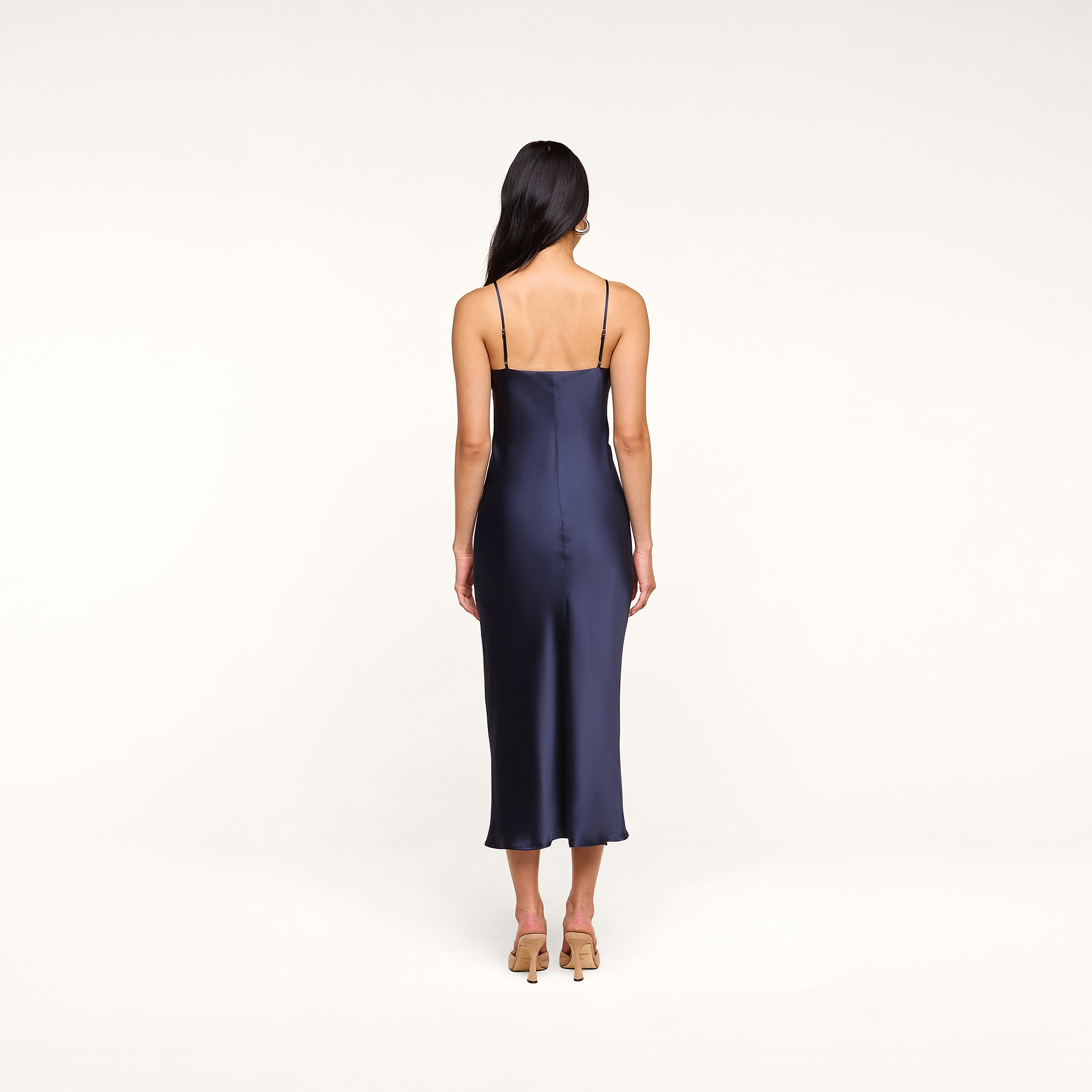 Wearing the Seridie Silk Slip Dress in navy blue, a woman with long dark hair stands facing away to reveal the elegant back and delicate spaghetti straps of this midi-length silhouette. Paired with beige heeled sandals, her look is effortlessly refined—ideal for a wedding guest.