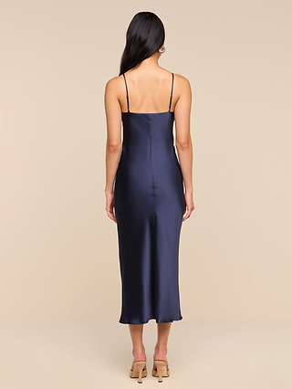 A woman with long dark hair wears the Seridie Silk Slip Dress in navy blue luxe satin, styled with beige heeled sandals, standing with her back to the camera against a plain beige background—an elegant look perfect for wedding guests.