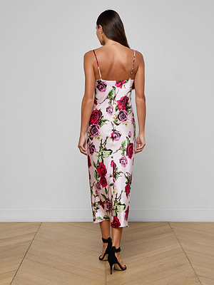A woman with long brown hair wears the Seridie Silk Slip Dress in pink with a rose print and black high-heeled sandals, standing on a wooden floor facing away from the camera.