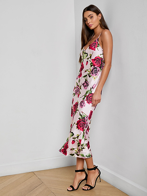 A woman stands against a white wall wearing the Seridie Silk Slip Dress, a silky, bias-cut midi featuring a pink and red rose print, paired with black strappy high-heeled sandals on a light wood floor.
