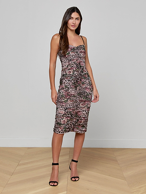 A woman with long hair and a neutral expression stands indoors on a wooden floor, wearing the Caprice Midi Dress in rose cheetah print with thin straps and black high-heeled sandals. A plain, light background highlights this figure-flattering dress.