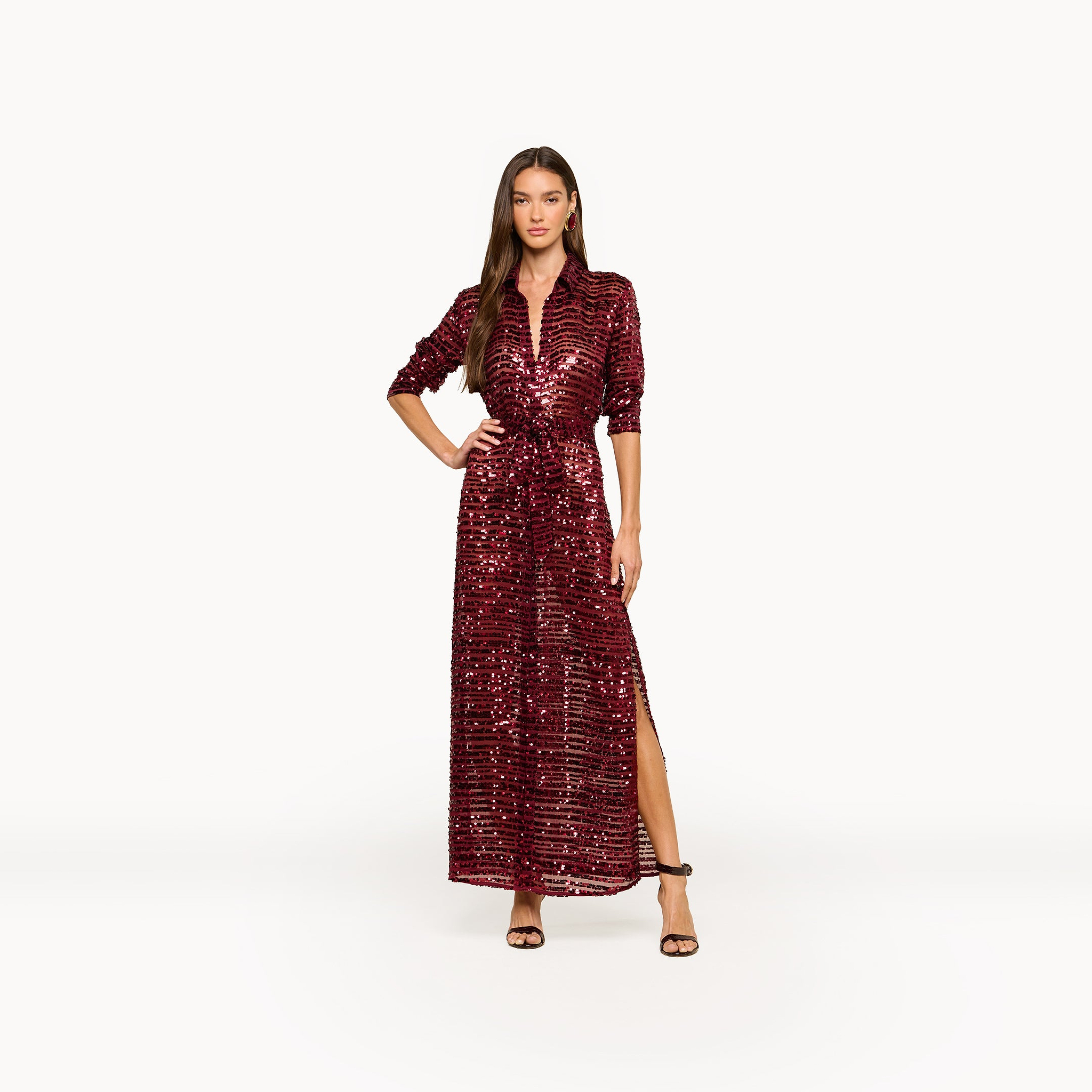 A woman poses in the Cameron Sequin Shirt Dress, a maxi-length red sequin dress featuring a slit and three-quarter sleeves. She stands with one hand on her hip, wearing brown ankle-strap heels against a plain white background.