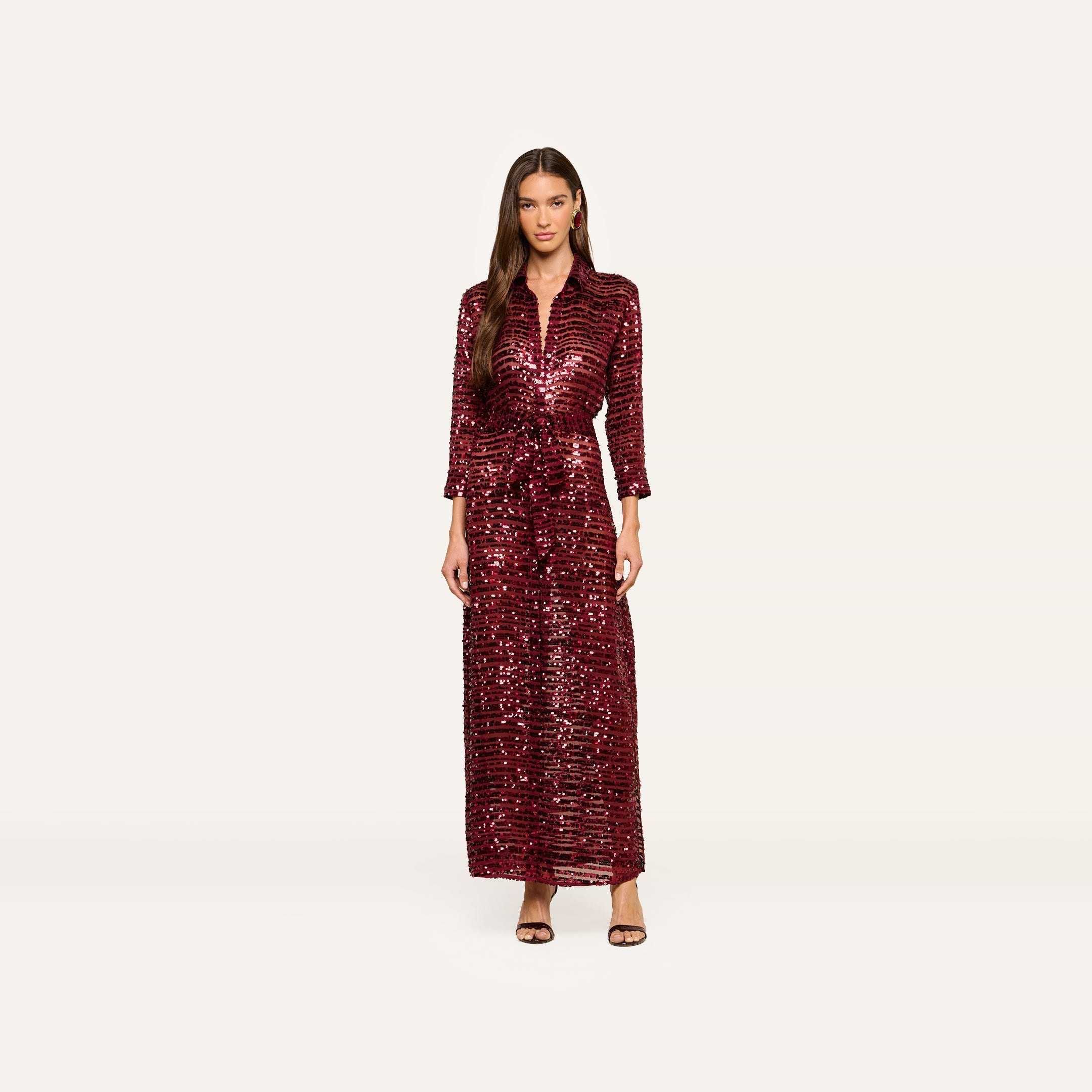 A woman stands against a plain white background wearing the Cameron Sequin Shirt Dress, a maxi-length, deep red sequin dress with long sleeves, paired with black strappy heels.