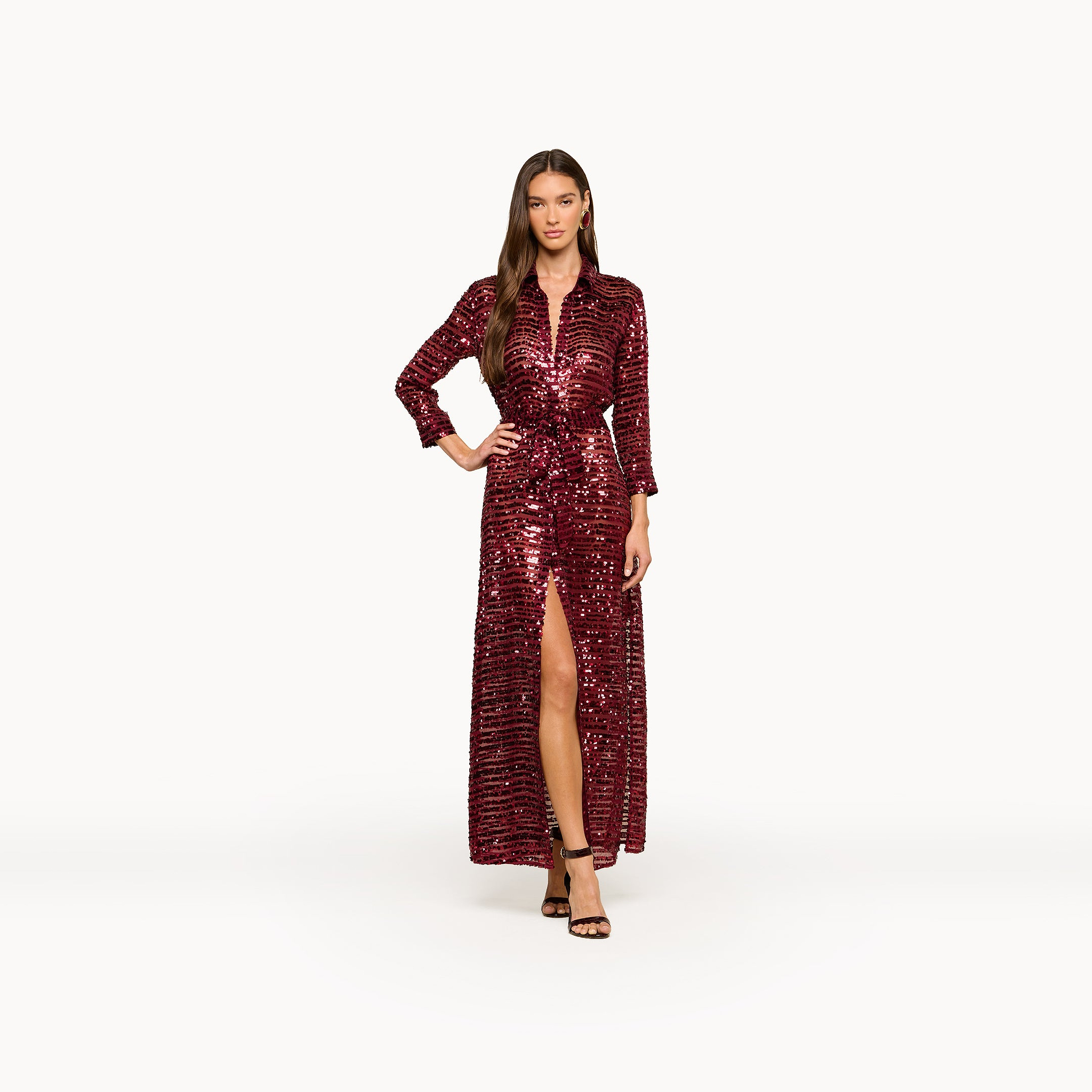 A woman stands facing forward against a plain white background, wearing the Cameron Sequin Shirt Dress—a maxi-length dress in deep red with sequined stripes and a front slit. She has long hair and pairs the look with black strappy heels.