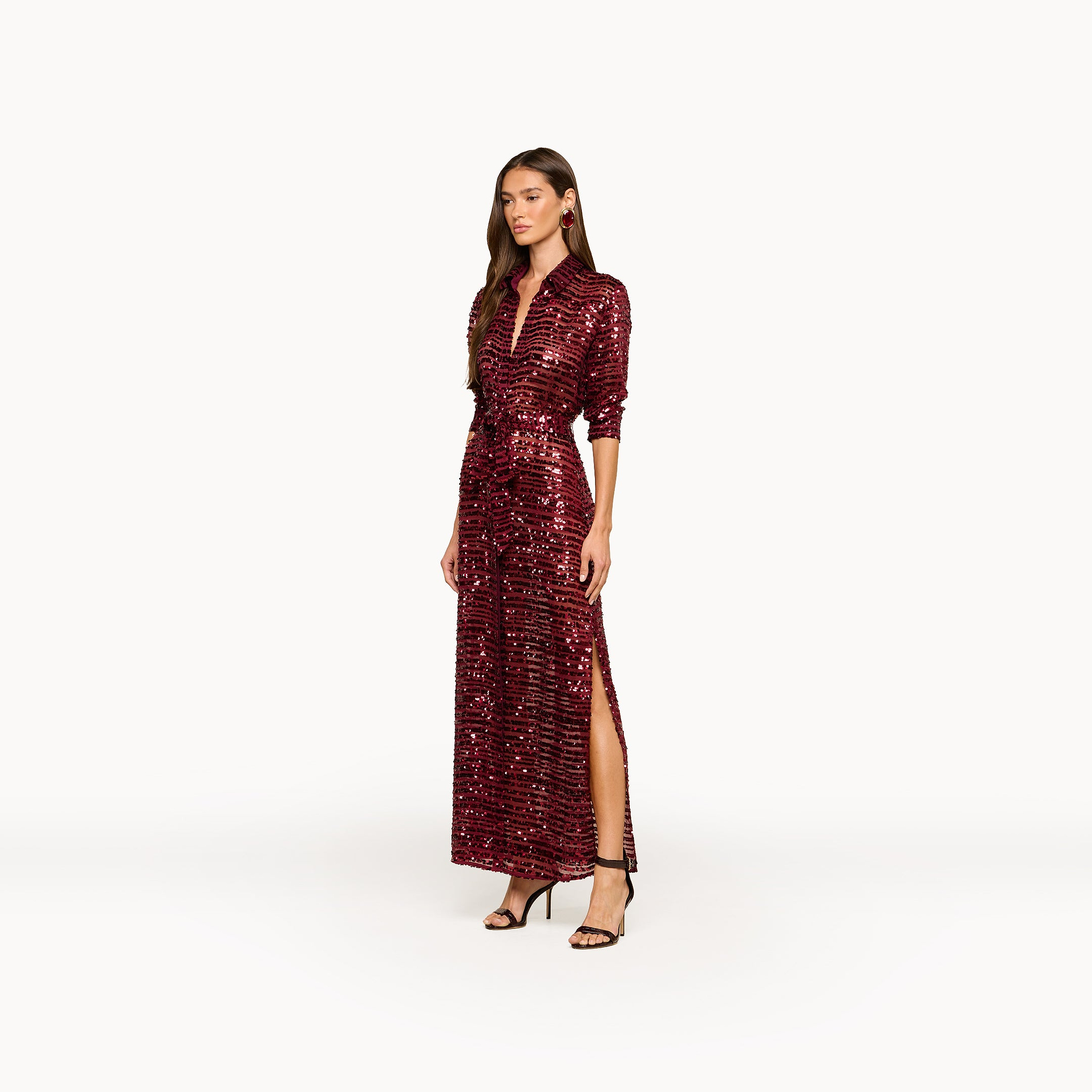 A woman wearing the Cameron Sequin Shirt Dress—a long, sparkling burgundy dress with sequined stripes, maxi length and a high slit, long sleeves—paired with black heels and standing against a plain white background.