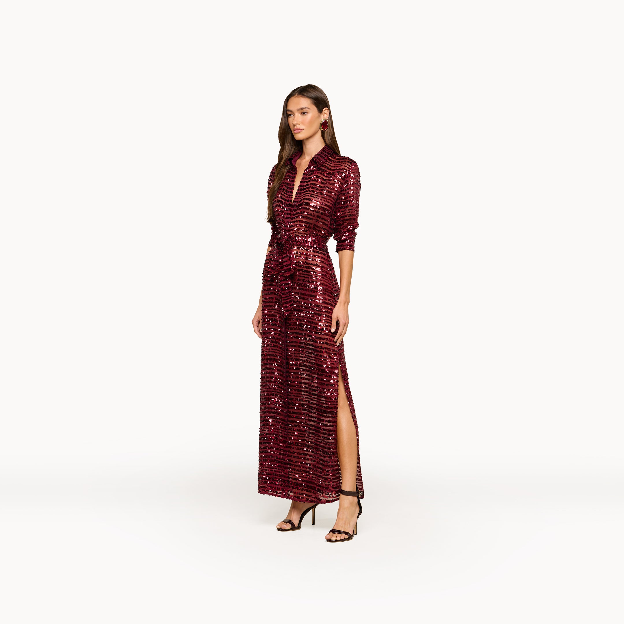 A woman wearing the Cameron Sequin Shirt Dress—a long, sparkling burgundy dress with sequined stripes, maxi length and a high slit, long sleeves—paired with black heels and standing against a plain white background.