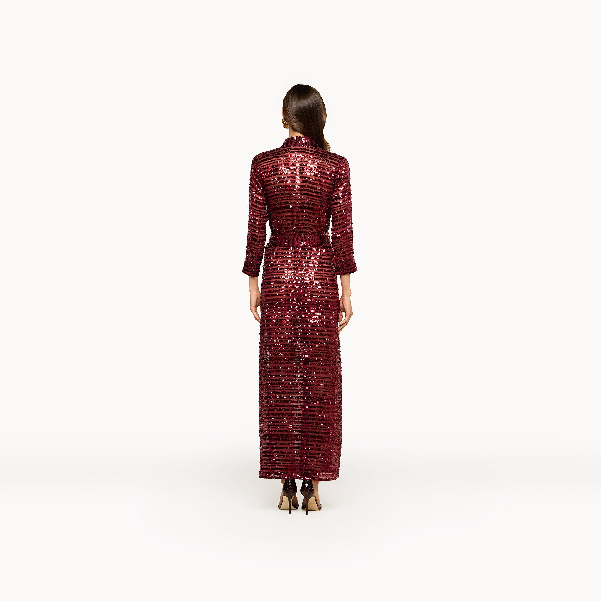 A woman stands facing away, wearing the maxi-length, long-sleeved Cameron Sequin Shirt Dress in red with black heels against a plain white background.