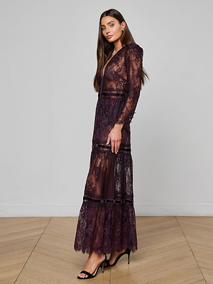 A woman with long brown hair wears the Poe Lace Maxi Dress—a sheer, dark purple lace gown with velvet trim and tiered layers—paired with black high-heeled sandals, standing on a wooden floor against a plain light gray wall.