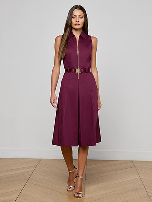 A woman wears the L'AGENCE Kylo Poplin Shirt Dress—sleeveless, knee-length, maroon with a zip-up front and belted waist—paired with metallic high-heeled sandals, standing on a wooden floor against a light gray backdrop.