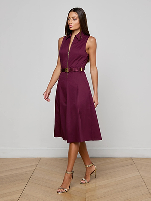 A woman wears the L'AGENCE Kylo Poplin Shirt Dress, a sleeveless maroon cotton design with a belted waist and gold buckle, paired with gold open-toe heels on a light wooden floor against a plain light gray wall.