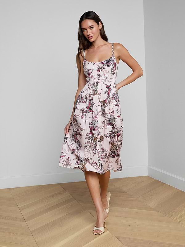 A woman smiles as she poses against a plain white background, wearing the Reanne Midi Dress— a white linen dress featuring a floral pattern and thin straps.