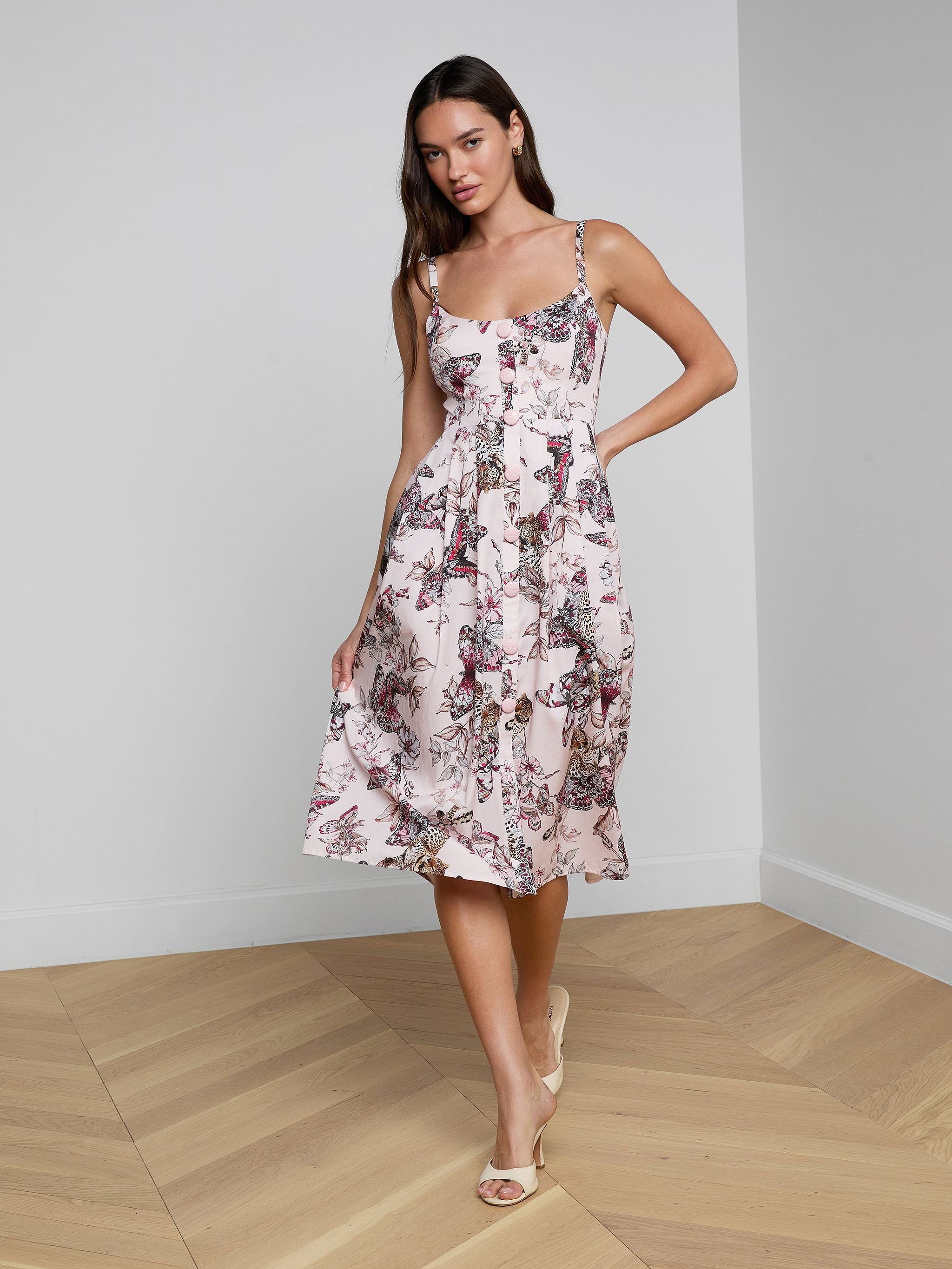 A woman smiles as she poses against a plain white background, wearing the Reanne Midi Dress— a white linen dress featuring a floral pattern and thin straps.