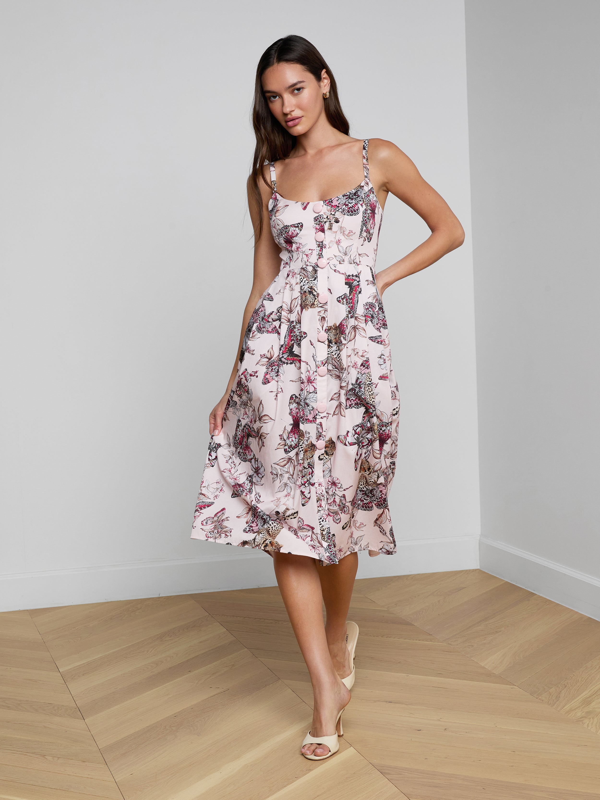 A woman smiles as she poses against a plain white background, wearing the Reanne Midi Dress— a white linen dress featuring a floral pattern and thin straps.