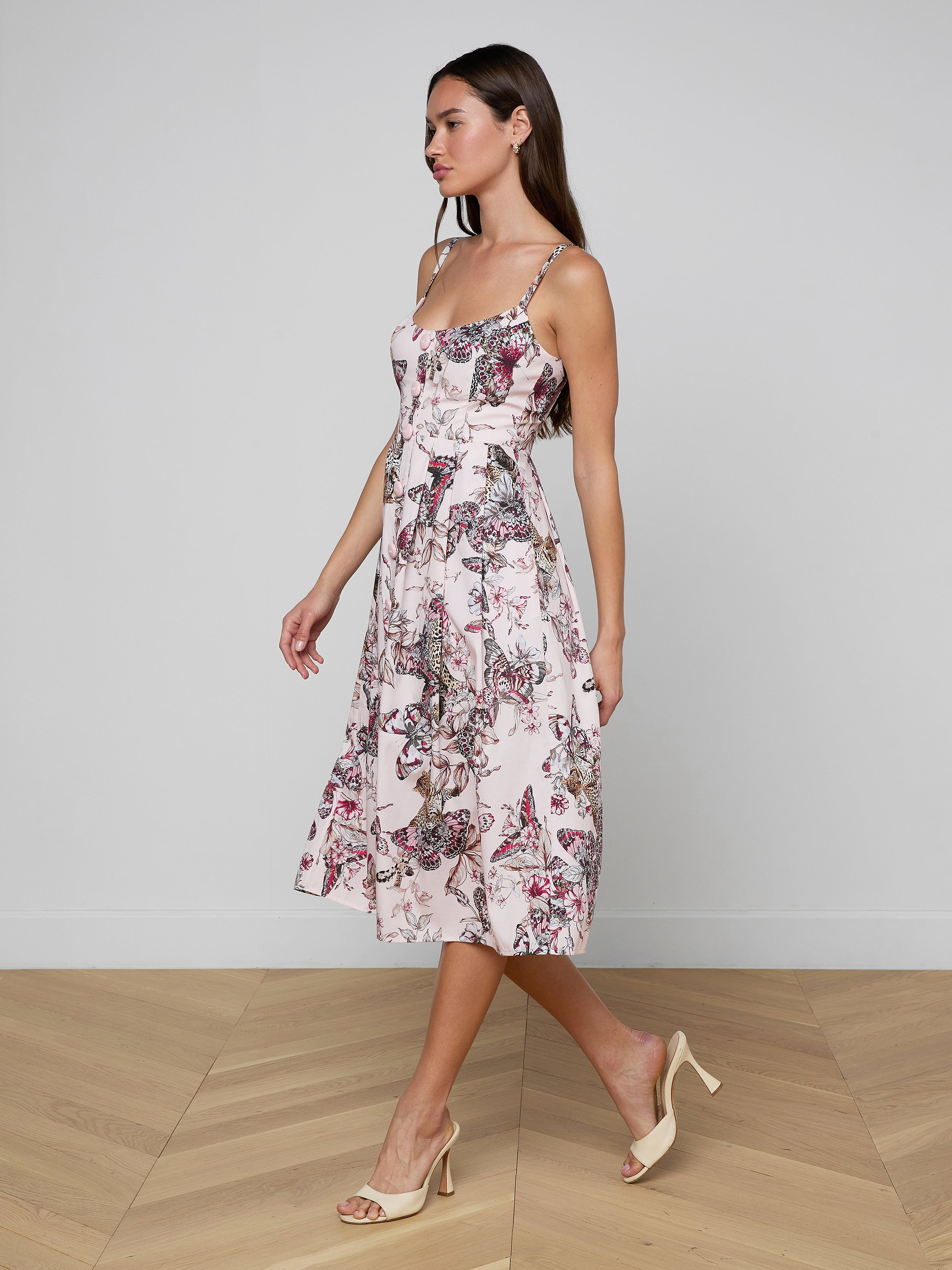 A woman in the Reanne Midi Dress, a light, sleeveless butterfly jungle print outfit with a floral pattern, paired with beige heels, walks to the left against a plain white background.