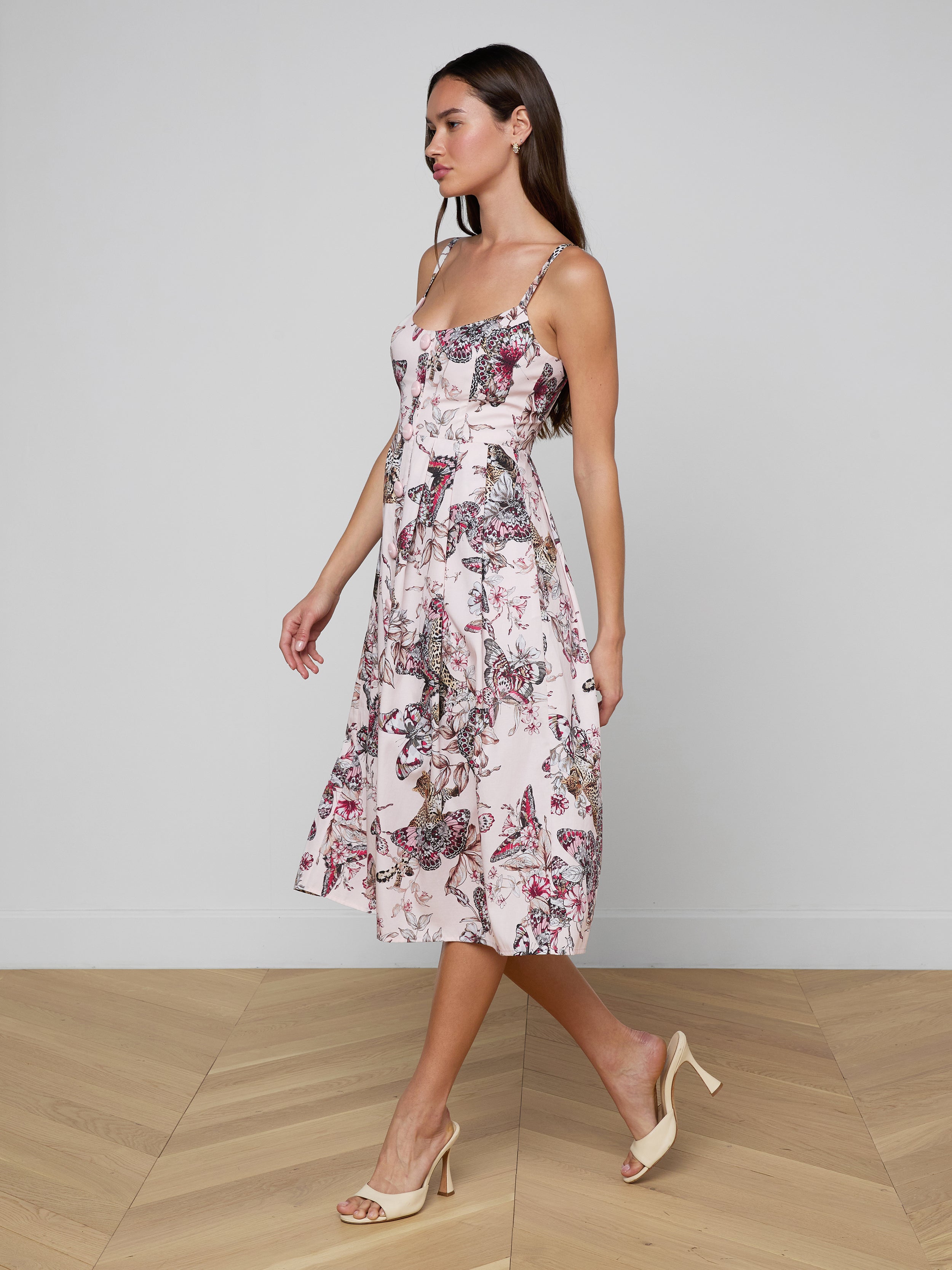 A woman in the Reanne Midi Dress, a light, sleeveless butterfly jungle print outfit with a floral pattern, paired with beige heels, walks to the left against a plain white background.