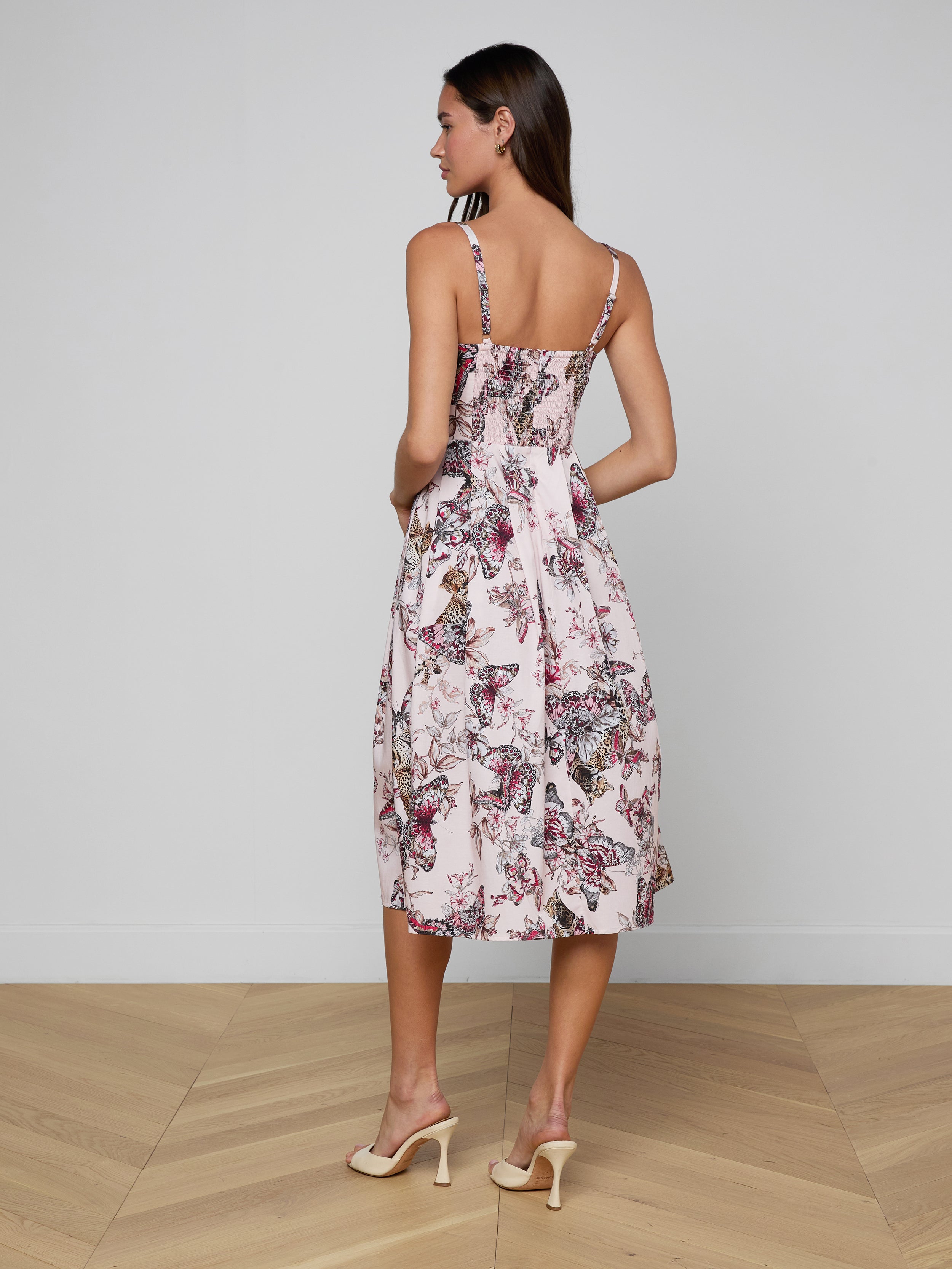 A woman stands facing away, wearing the Reanne Midi Dress—with a butterfly jungle print, thin straps, and midi length—paired with beige heels against a plain white background.