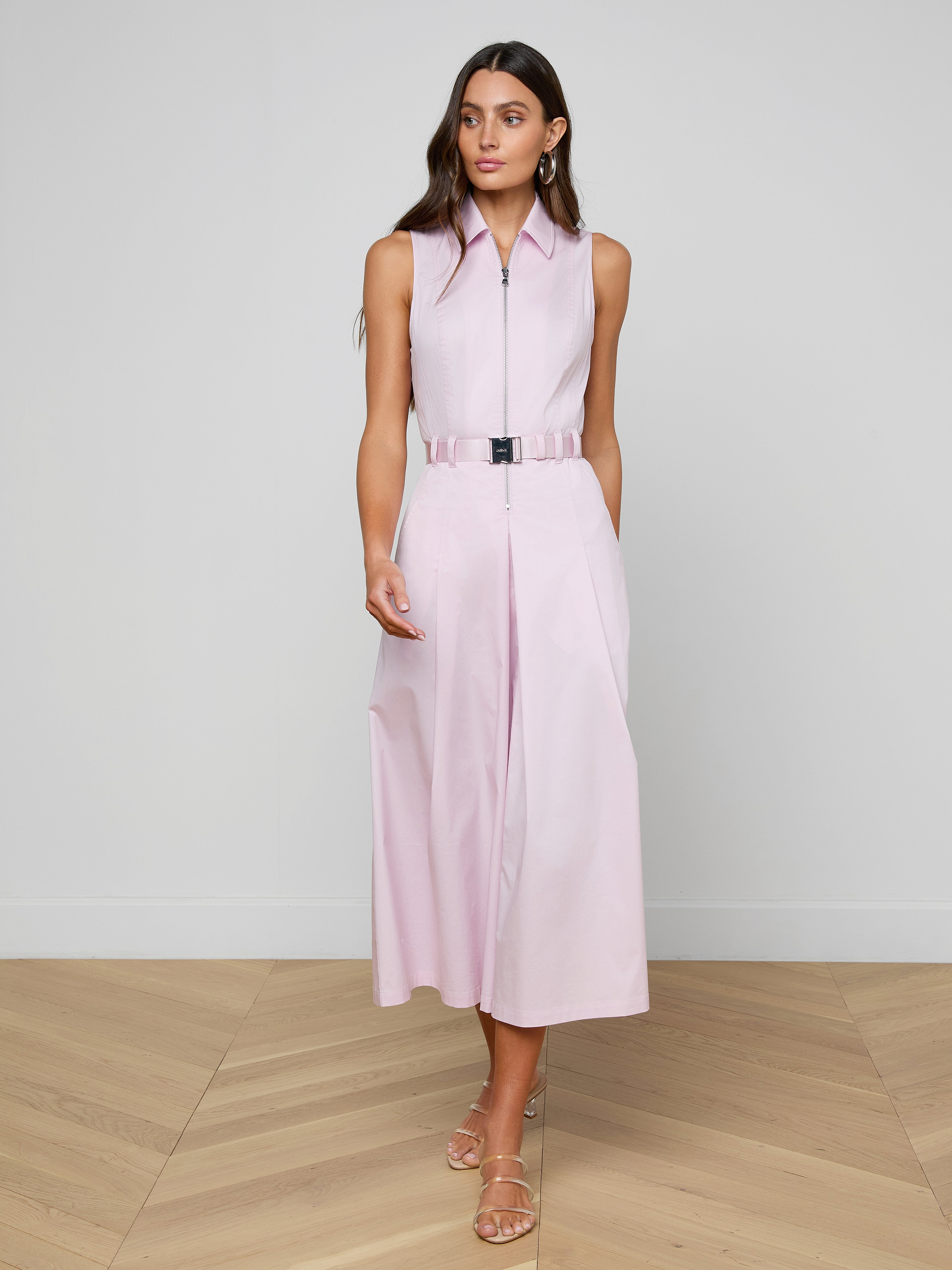 A woman wearing the Kolton Dress in sleeveless pastel lilac, styled with a statement belt and sandals, stands against a plain white background, gazing slightly to her right.