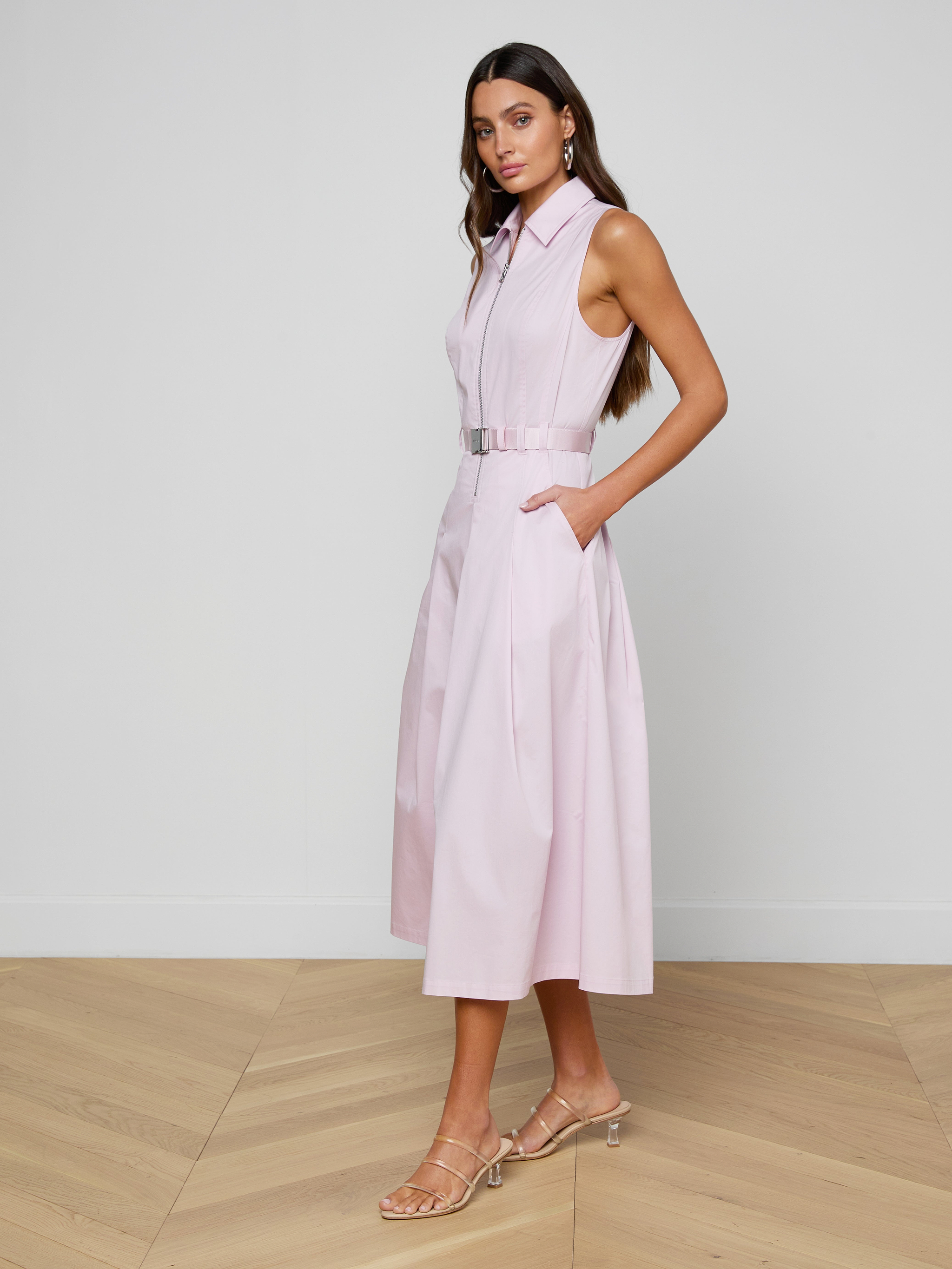 A woman stands against a white background wearing the Kolton Dress—a sleeveless, belted pastel lilac dress with pockets—paired with heeled sandals.