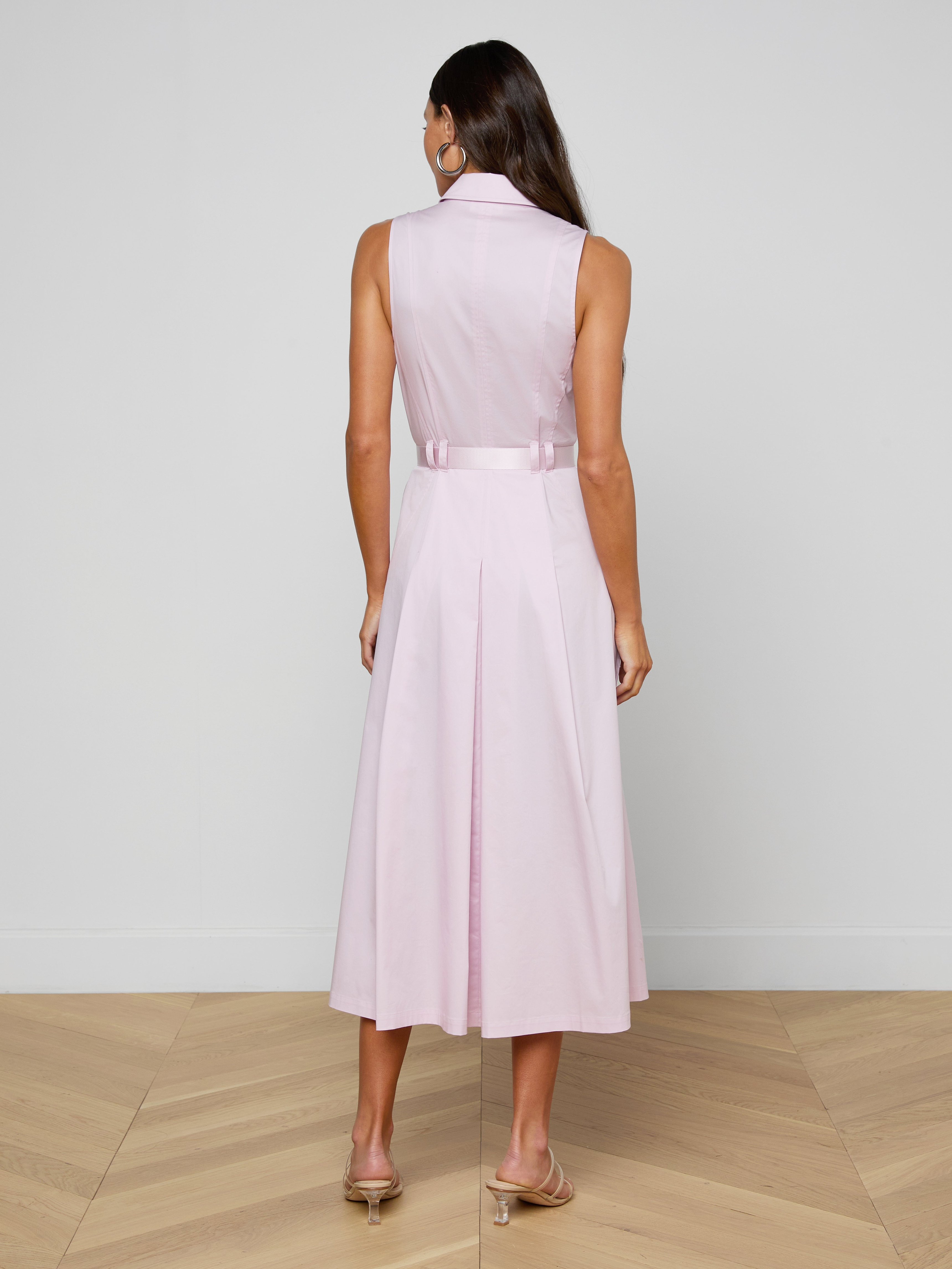 A woman stands with her back to the camera, modeling the sleeveless Kolton Dress in pastel lilac, accented with a statement belt and paired with beige heels against a plain white backdrop.