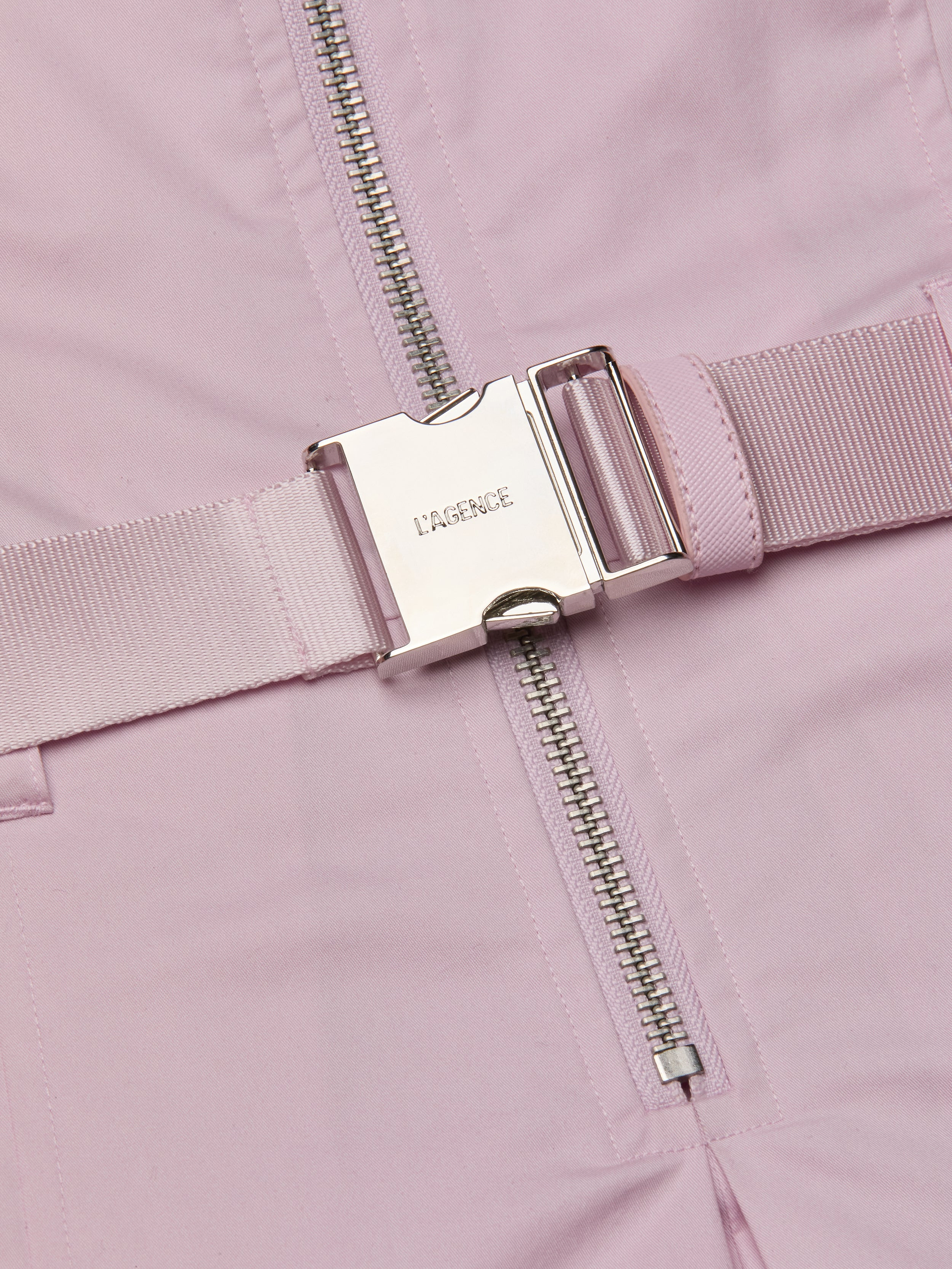 A close-up of the Kolton Dress features a pastel lilac statement belt with a shiny silver buckle engraved with LAGENCE, situated next to a silver zipper on matching pink fabric.