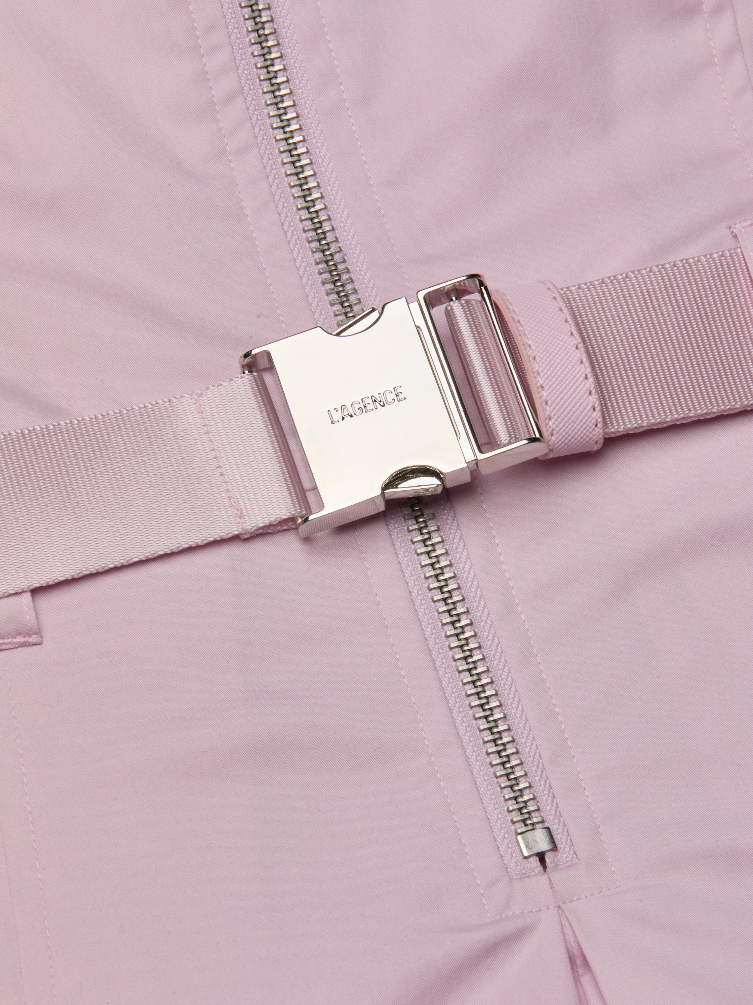 A close-up of the Kolton Dress features a pastel lilac statement belt with a shiny silver buckle engraved with LAGENCE, situated next to a silver zipper on matching pink fabric.