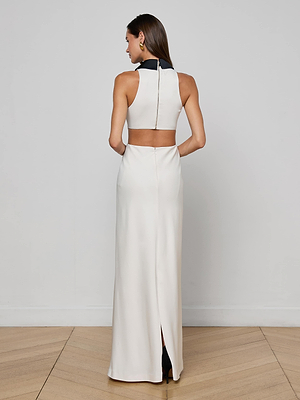 A person stands on a wood floor facing away, wearing the Rossi Cutout Gown by L’AGENCE—a sleeveless white maxi dress with a black collar, exposed back, and back slit—embodying minimalism against a plain white wall.