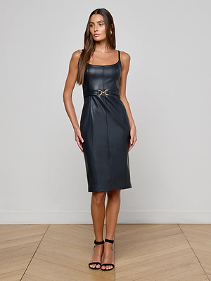 A woman poses indoors in the L'AGENCE Halia Vegan Leather Dress—a sleeveless, fitted black dress featuring a gold buckle belt—paired with black high-heeled sandals, against a plain light wall and wooden floor background.