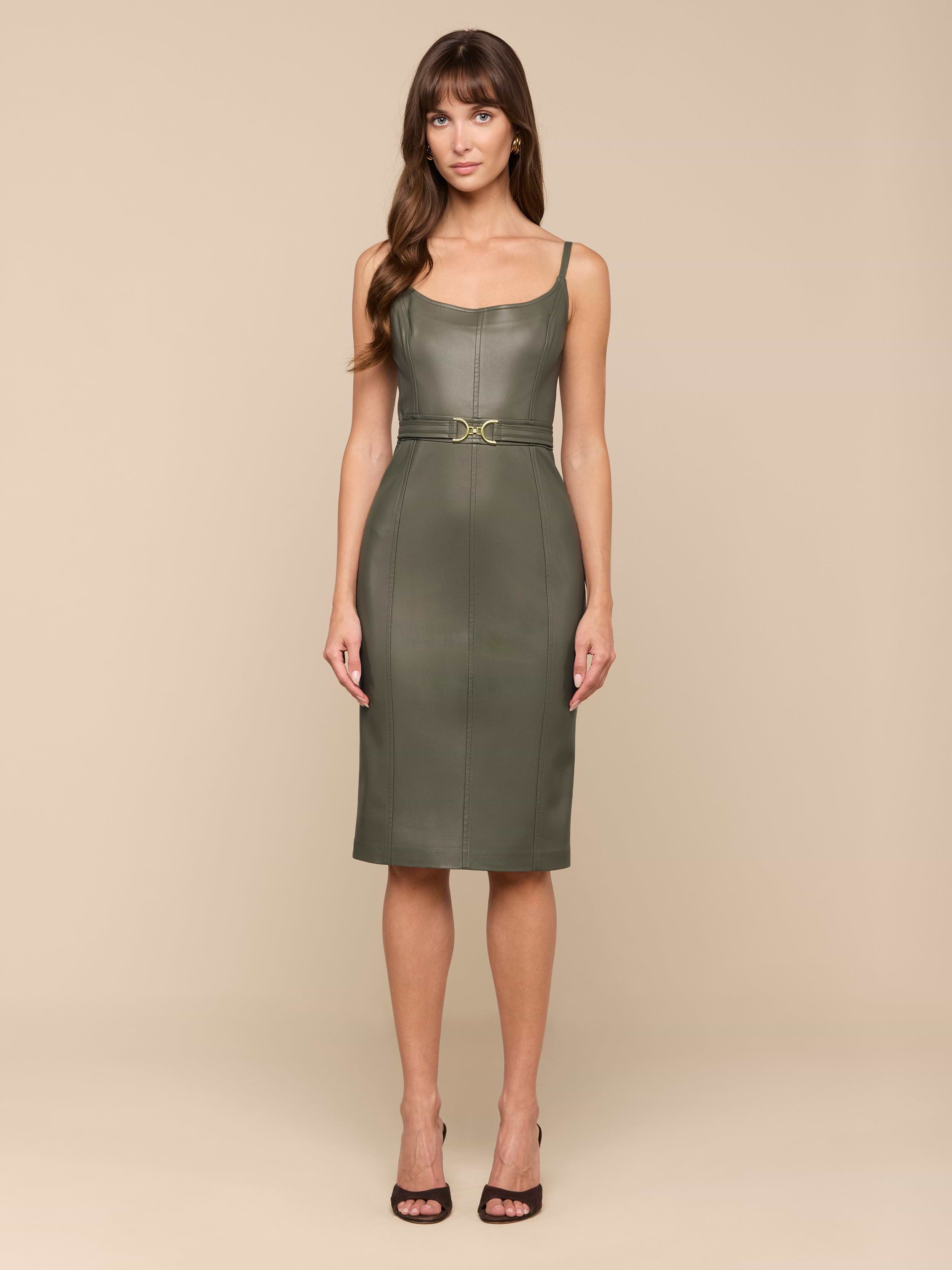 Une femme se tient debout sur un fond beige, de face, portant la robe Halia Vegan Leather Dress en smoky olive - une robe midi sans manches avec une taille ceinturée - associée à des sandales à talons noires.