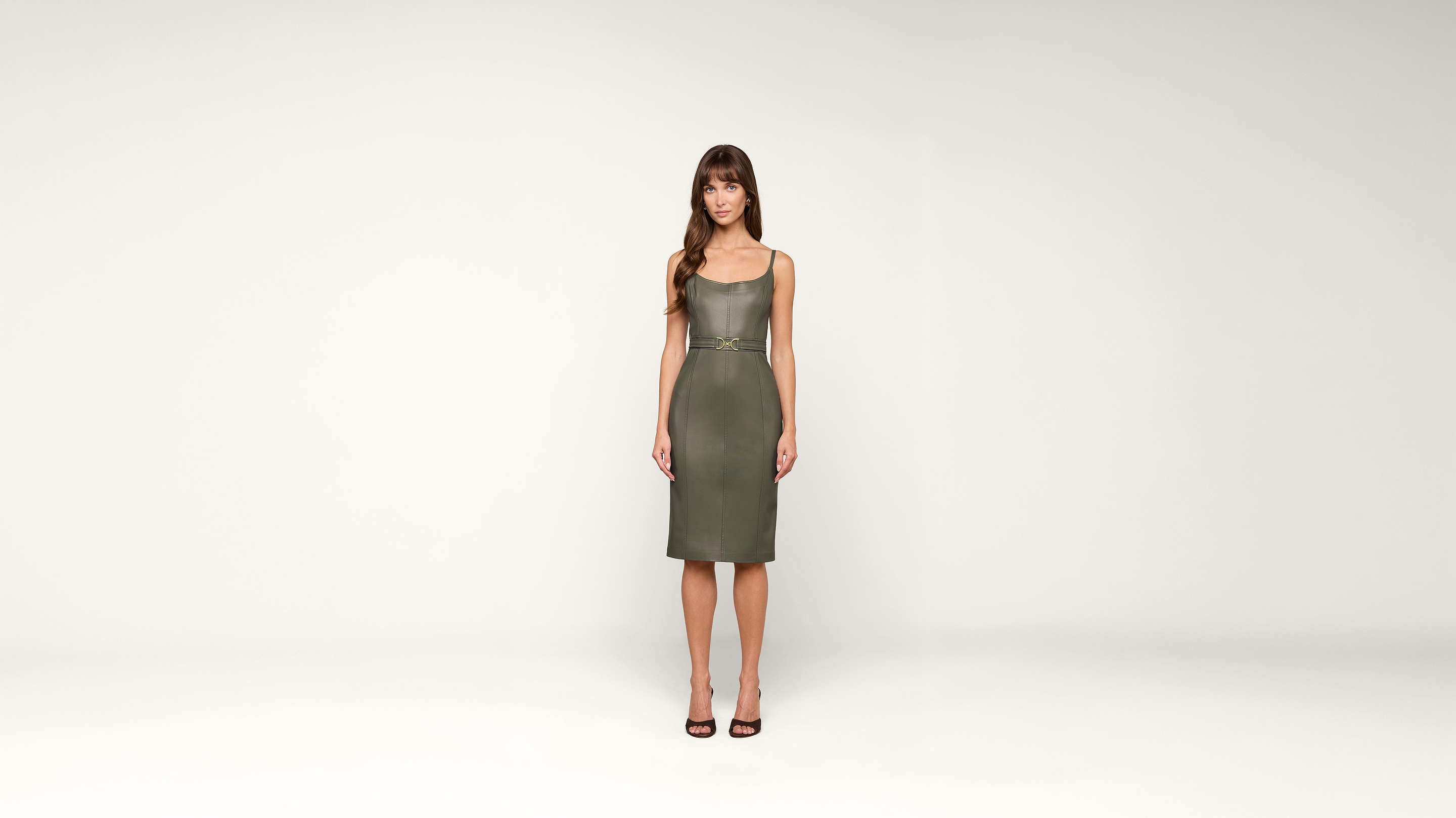 A woman with long brown hair stands facing forward, wearing a knee-length olive-green dress and black heels, against a plain white background.