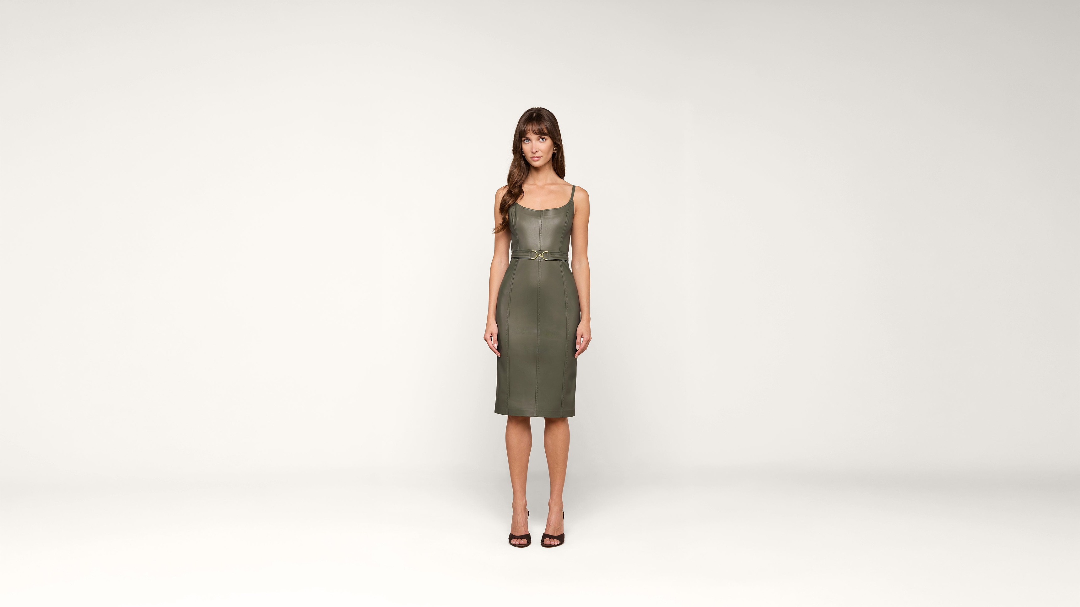A woman with long brown hair stands facing forward, wearing a knee-length olive-green dress and black heels, against a plain white background.
