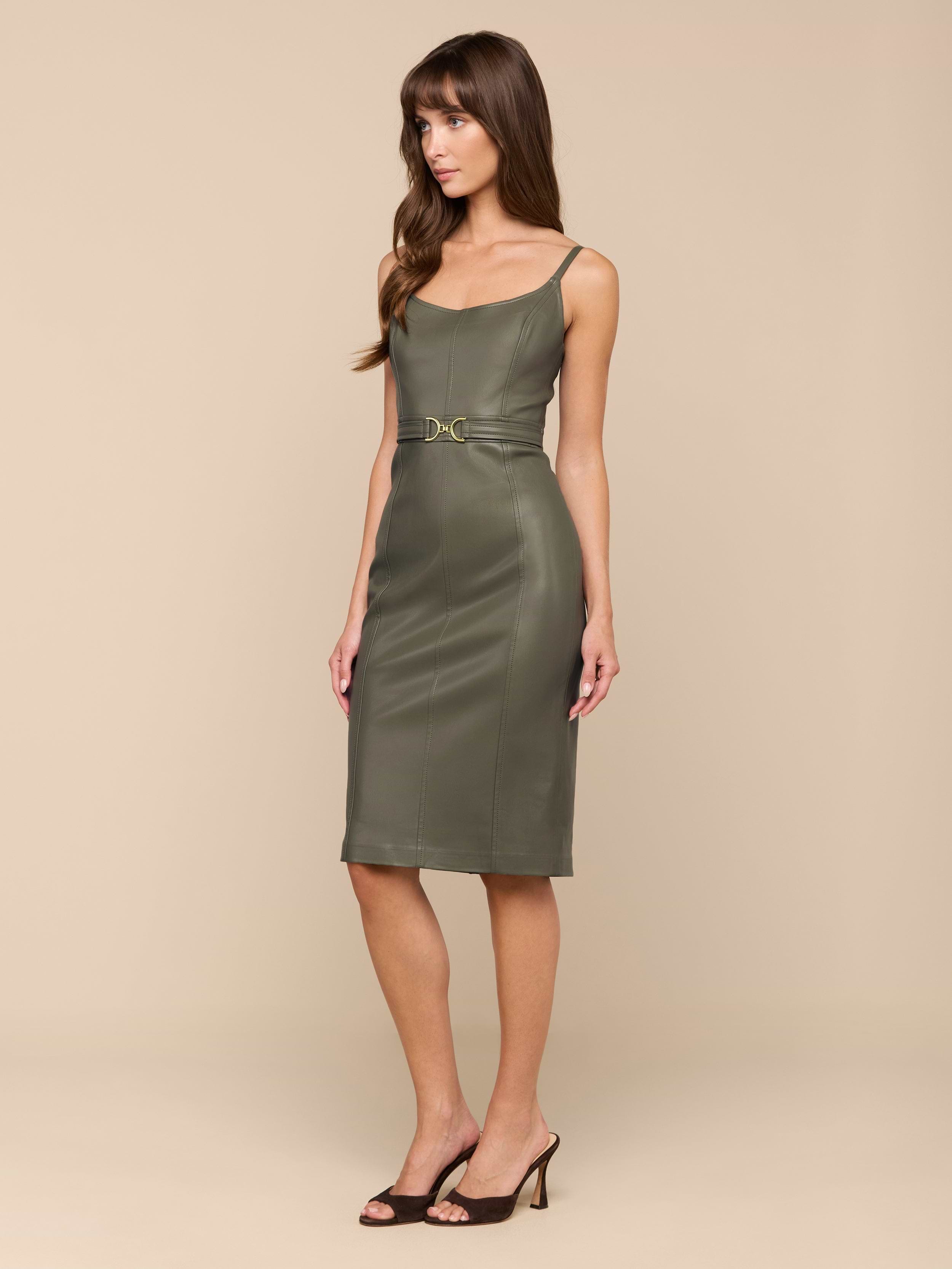 Une femme se tient debout sur un fond beige et porte la robe en cuir végétal Halia en olive fumé. La robe midi est dotée de fines bretelles et dune ceinture à la taille. Elle a de longs cheveux bruns et associe son look à des talons noirs à bout ouvert.