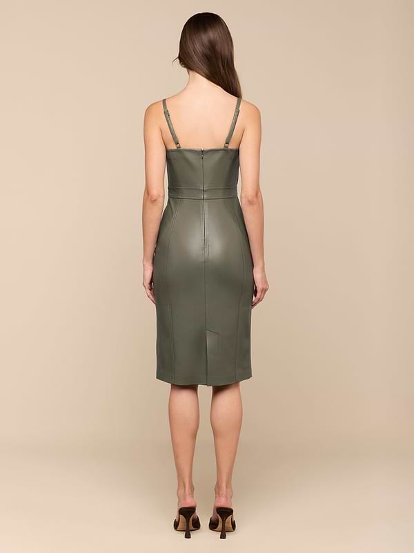 Une femme aux longs cheveux bruns se tient de face, portant la robe Halia Vegan Leather Dress en olive fumé avec de fines bretelles et des détails de couture dans le dos, ainsi que des sandales à talons brunes, sur un fond beige.
