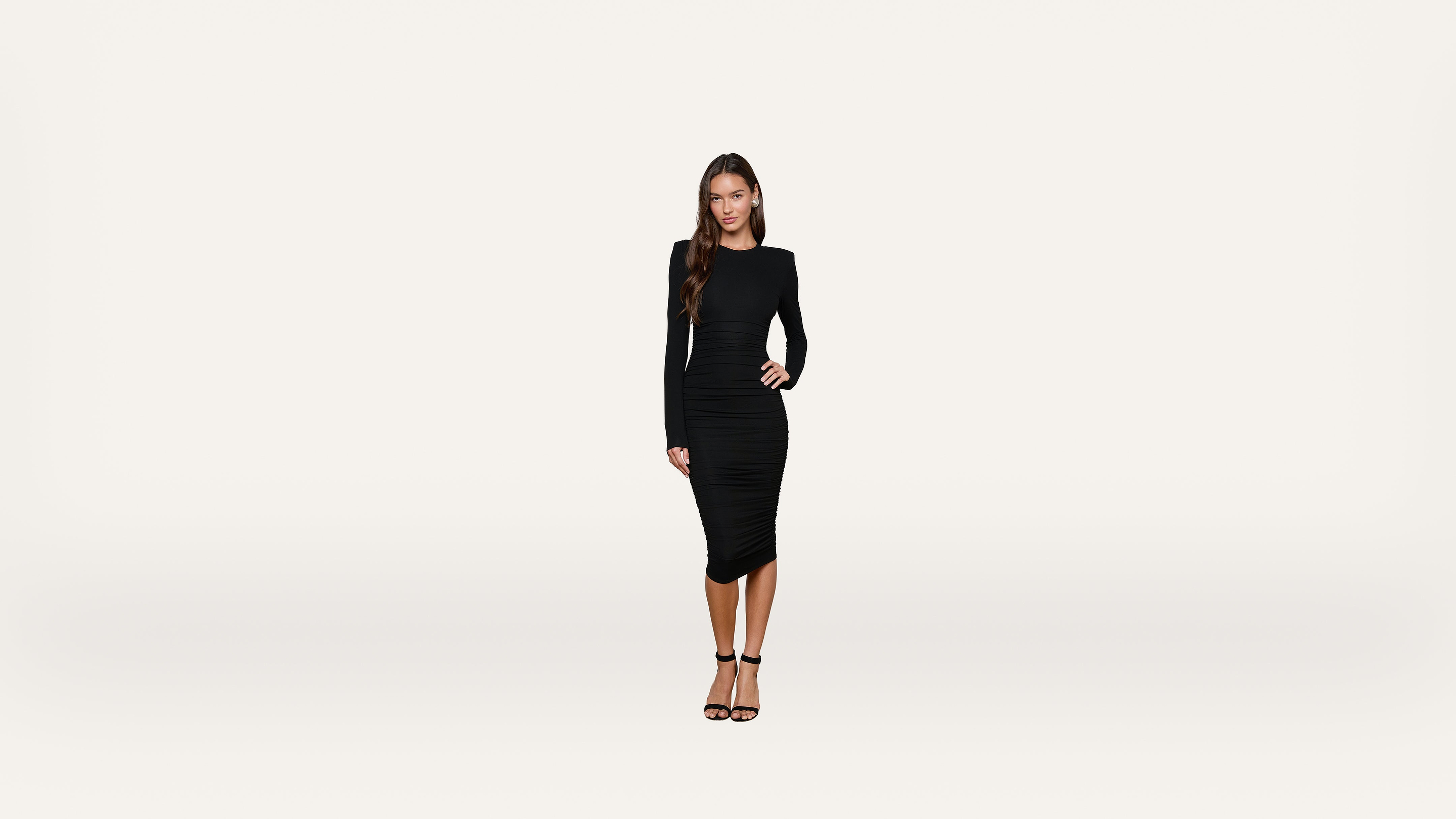 A woman with long hair wearing a fitted black dress and black heels stands against a plain white background, looking at the camera with a neutral expression.