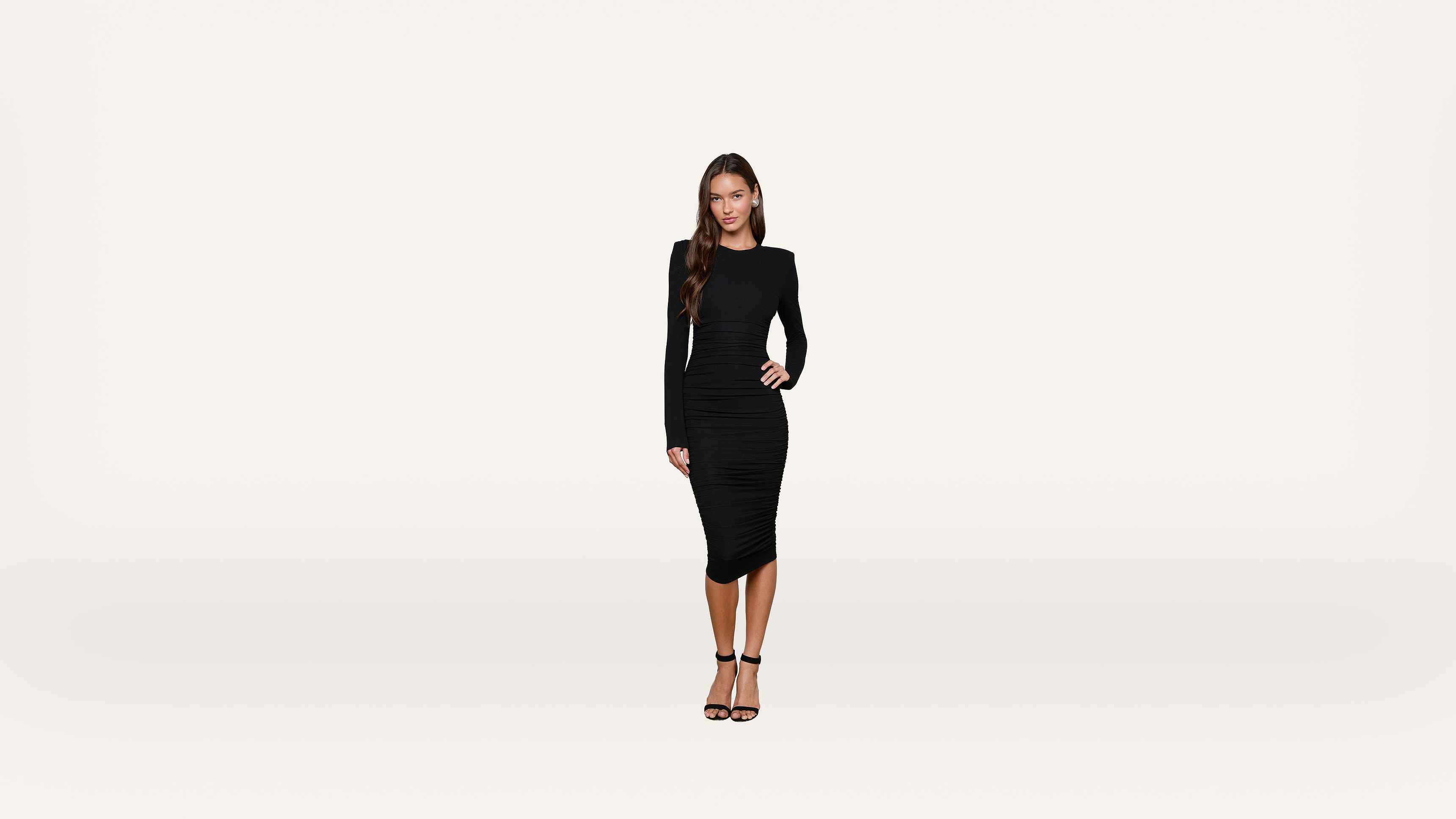 A woman with long hair wearing a fitted black dress and black heels stands against a plain white background, looking at the camera with a neutral expression.