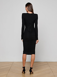 A woman stands facing away, wearing the Paityn Shirred Dress—a long-sleeved, fitted black dress crafted from stretch jersey with subtle ruching—paired with black high-heeled shoes, against a plain white background.