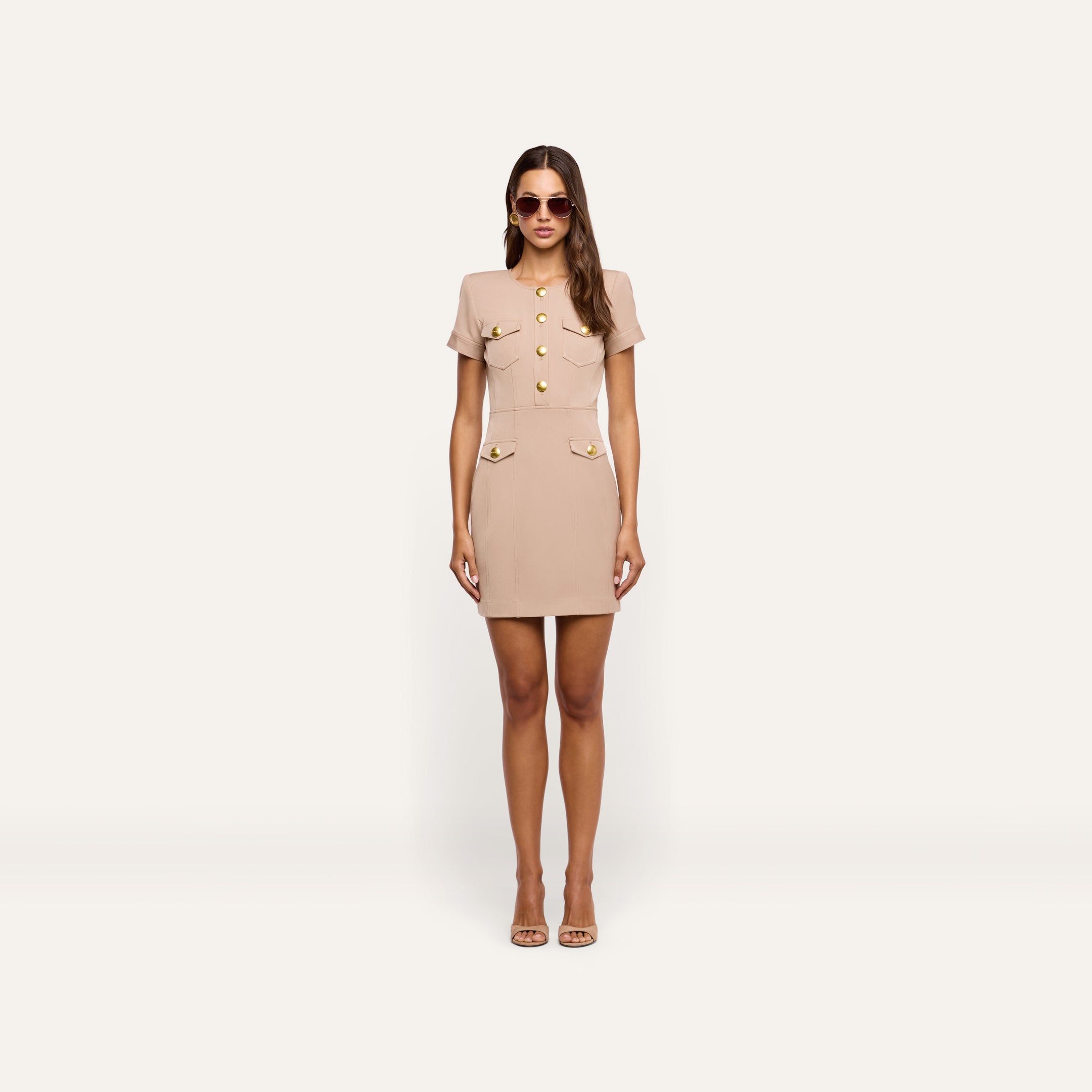 A woman wearing a tan Cori Mini Dress with gold-tone hardware and two front pockets, paired with sunglasses and open-toe heels.