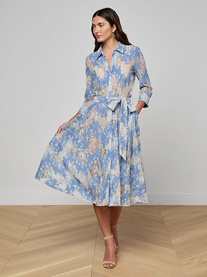 A woman poses indoors in the Ralphie Lace Midi Dress—light blue, long-sleeve, floral with a waist tie—paired with nude heeled sandals. She stands on a light wood floor against a plain white wall.