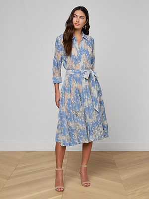 A woman stands on a wooden floor wearing the Ralphie Lace Midi Dress, a light blue, floral-patterned shirt dress with a matching waist tie and long sleeves, styled with beige heeled sandals.