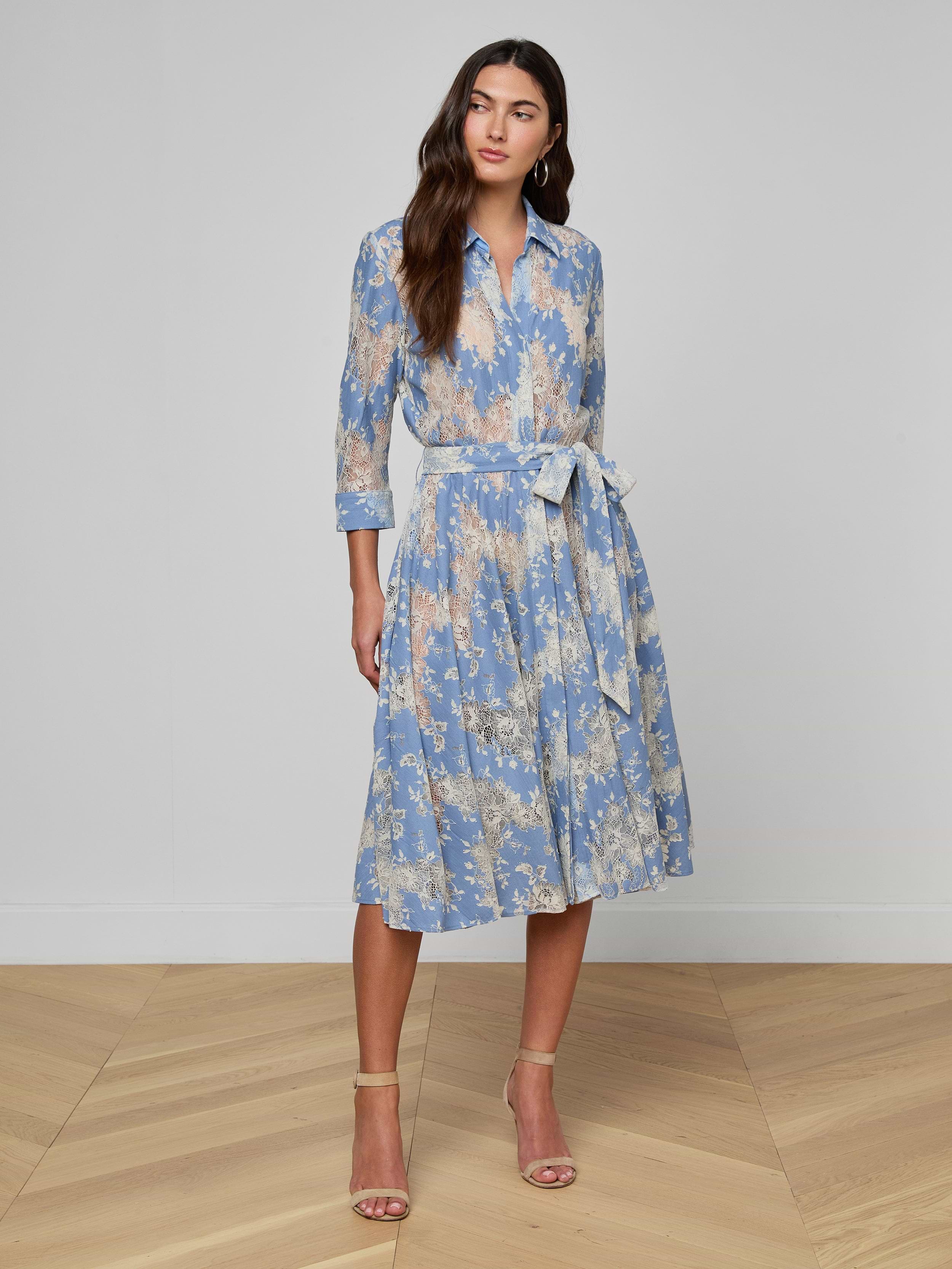 Woman wearing the Ralphie Lace Midi Dress in light blue, featuring long sleeves, a knee-length silhouette, floral lace detailing, and a tied waist; styled with beige heels against a plain white background.
