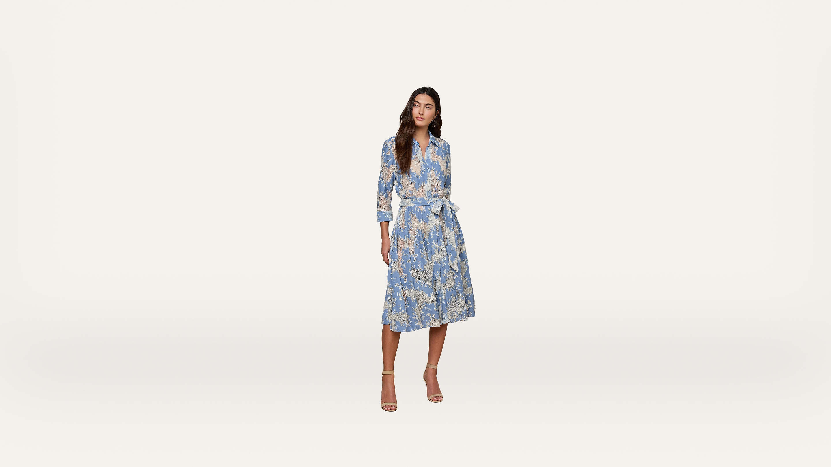 A woman stands wearing a light blue and yellow floral dress with long sleeves and a matching belt, paired with beige high-heeled sandals, against a plain white background.