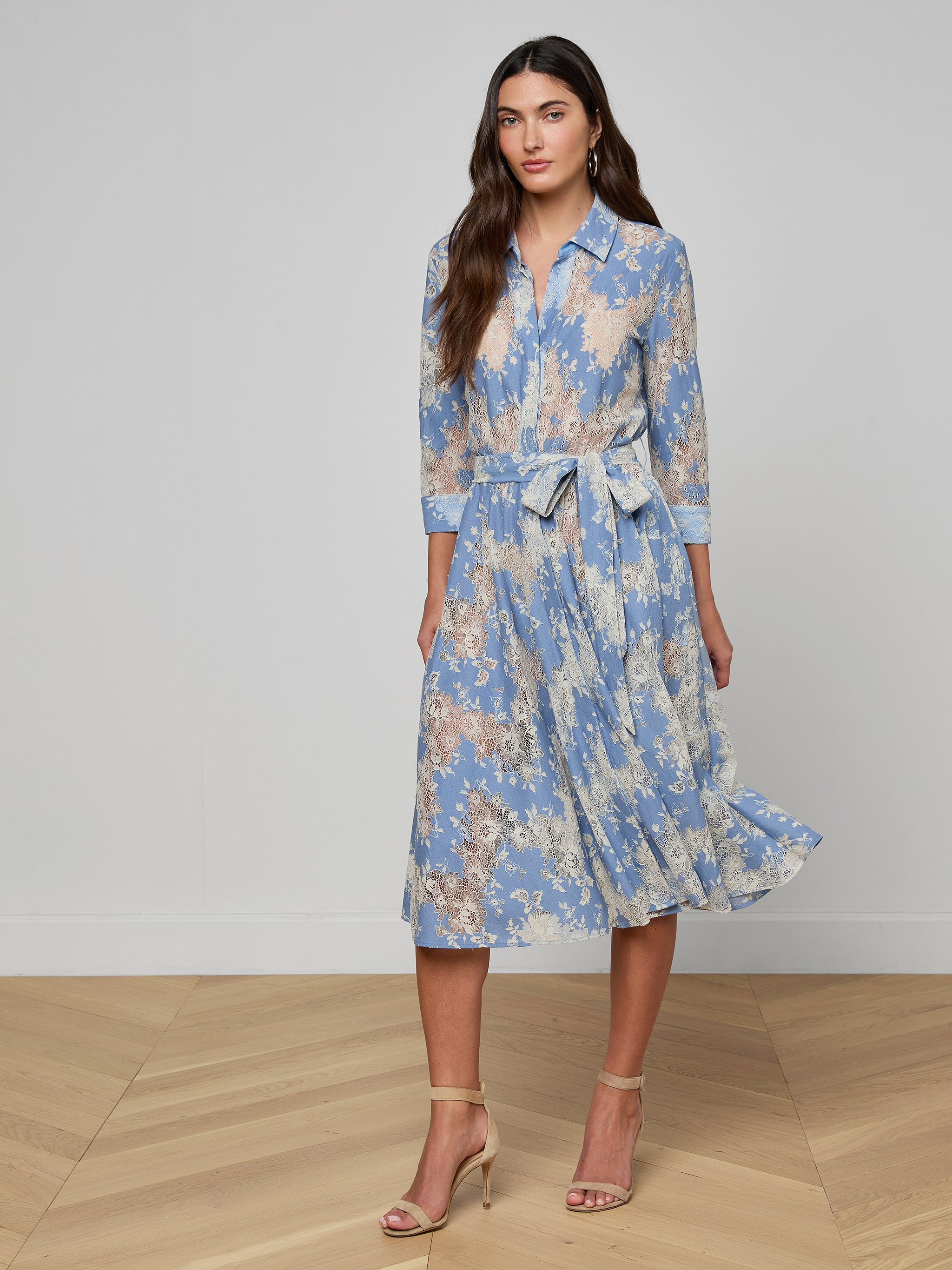 A woman wearing a light blue and beige floral Ralphie Lace Midi Dress with long sleeves and a tie belt, paired with beige high-heeled sandals, stands against a plain white background.