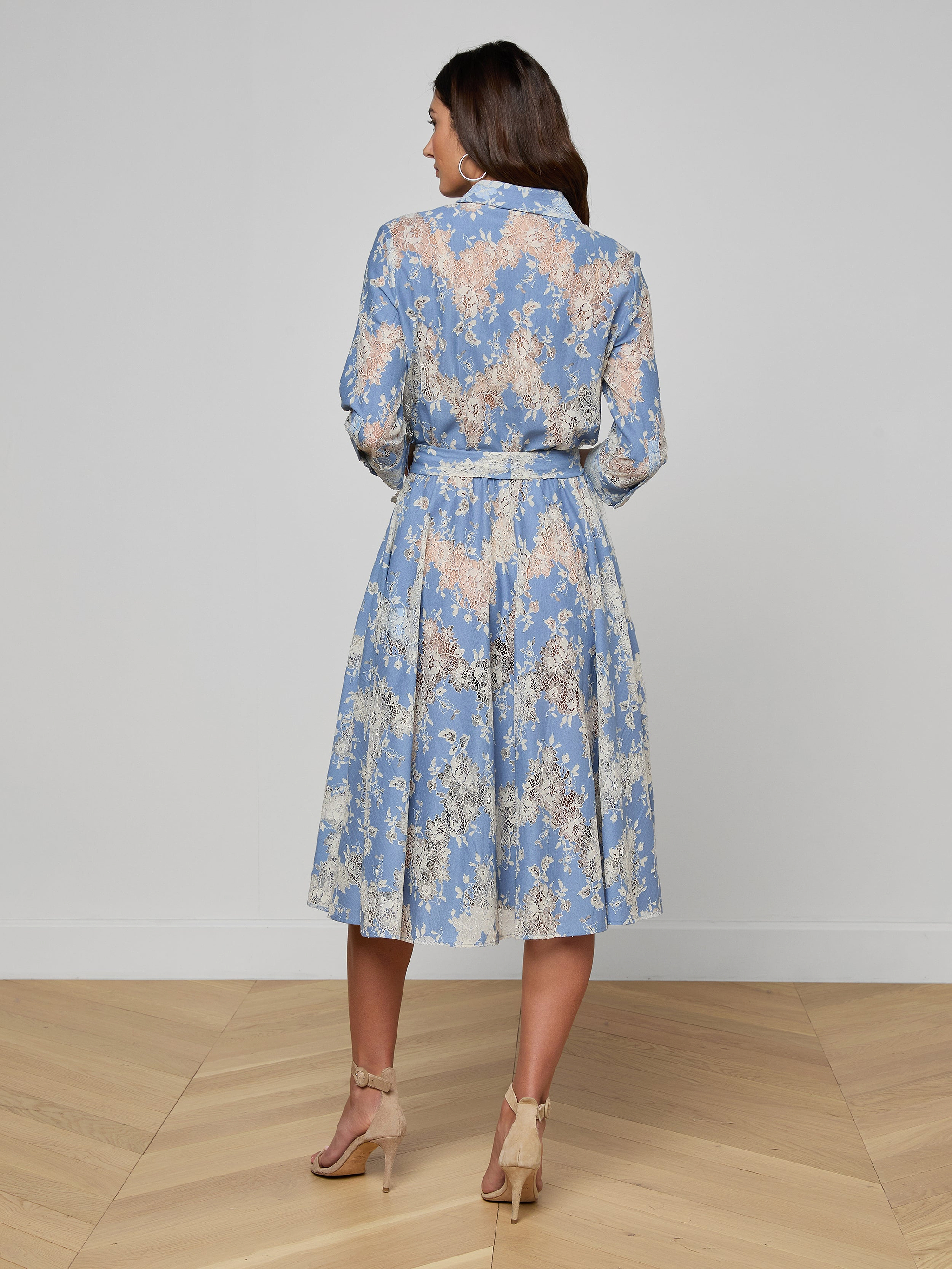 Woman standing in heels, facing away, wearing a light blue Ralphie Lace Midi Dress featuring long sleeves, a belted waist, and a mid-length skirt against a plain white background.