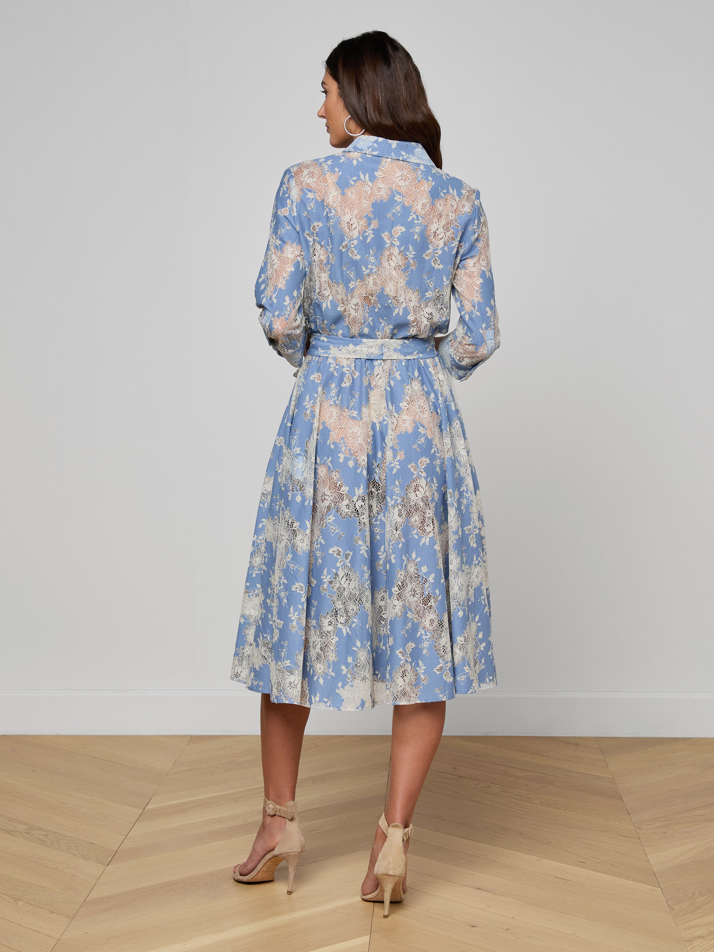 Woman standing in heels, facing away, wearing a light blue Ralphie Lace Midi Dress featuring long sleeves, a belted waist, and a mid-length skirt against a plain white background.