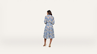 A woman stands facing away, wearing a light blue, long-sleeved, floral dress with a belted waist and beige heels, against a plain white background.