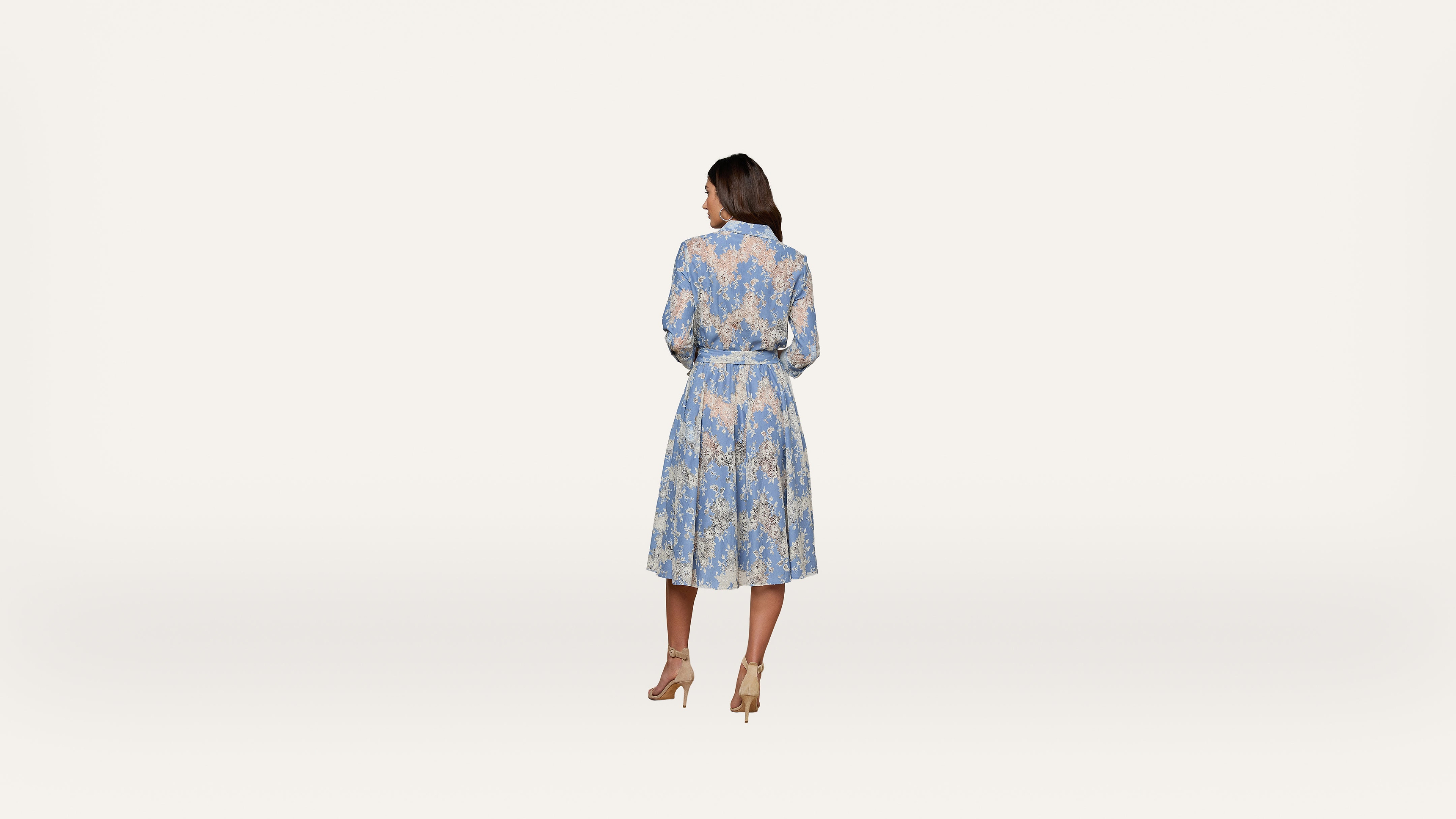 A woman stands facing away, wearing a light blue, long-sleeved, floral dress with a belted waist and beige heels, against a plain white background.