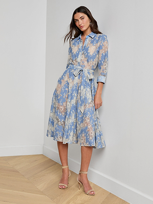 A woman stands indoors wearing the Ralphie Lace Midi Dress, a light blue long-sleeve shirt dress with a waist tie, paired with beige high-heeled sandals. She leans against a white wall on a wooden floor.