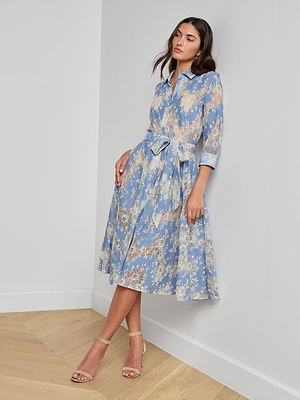 A woman wearing the Ralphie Lace Midi Dress—a light blue, floral-patterned shirt dress with a waist tie and three-quarter sleeves—stands against a white wall in beige heeled sandals, looking slightly to the side.