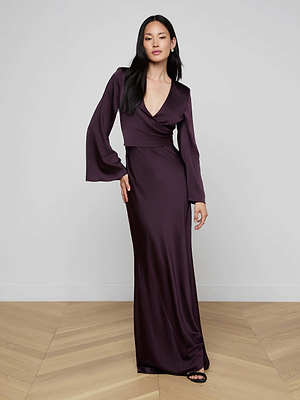 A woman with long, straight black hair poses indoors on a wooden floor, wearing the Manon Satin Gown—a long-sleeved, deep purple wrap dress with a plunging neckline and a floor-length skirt, her hand resting on her hip.