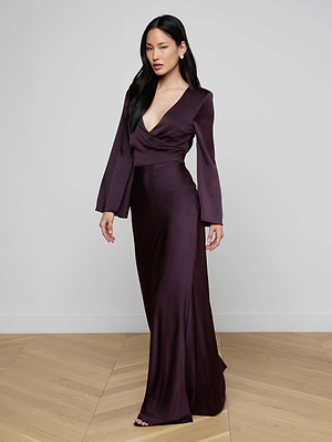 A woman with long dark hair stands indoors in the Manon Satin Gown, featuring a rich purple shade, floor-sweeping silhouette, and long sleeves. She poses against a plain light gray wall with a wooden floor.