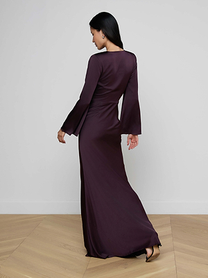 A woman with long dark hair wears the Manon Satin Gown, a floor-length plum dress, standing on light wood flooring and glancing over her left shoulder away from the camera.